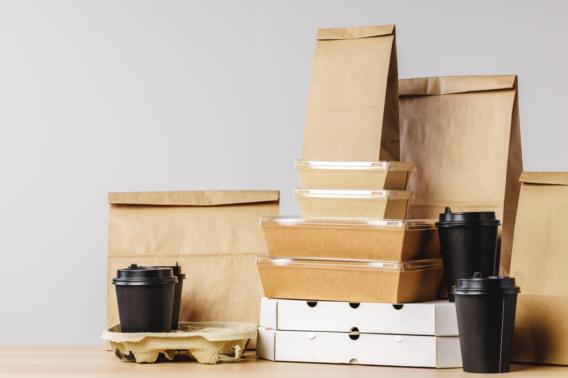 Stacks of brown paper bags, cardboard food containers, and black coffee cups with lids on a wooden surface against a neutral background.